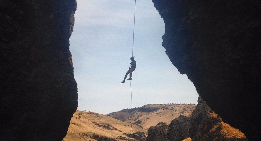 Wearing safety gear is suspended by ropes, midair, between two rock walls. 